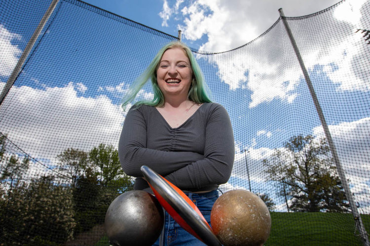 Picture is of a smiling young white woman with long green hair wearing a long-sleeved gray shirt with arms crossed. She is standing outside with track and field equipment under a blue sky with puffy white clouds.