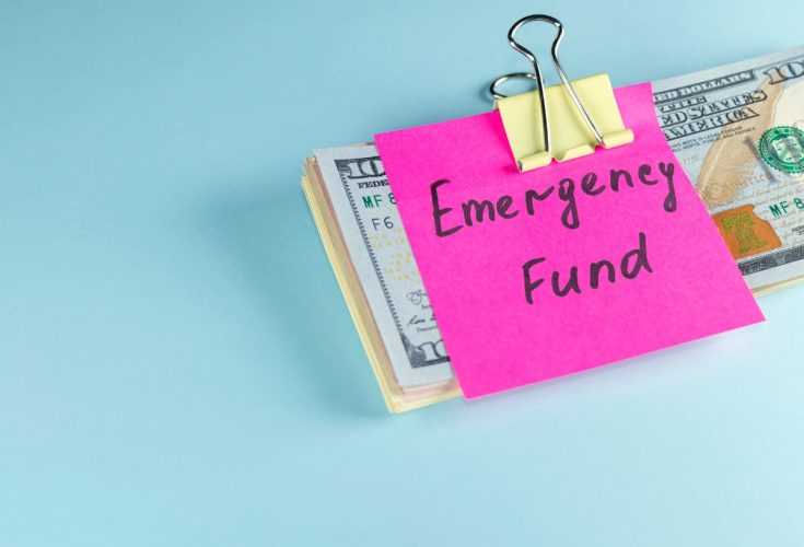 Aqua background with a small stack of US currency clipped together a Post-It Note on top reading "emergency fund."